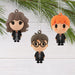 Three small hanging character ornaments shaped like Harry Potter characters are displayed against a white wood background with green pine branches at the top.