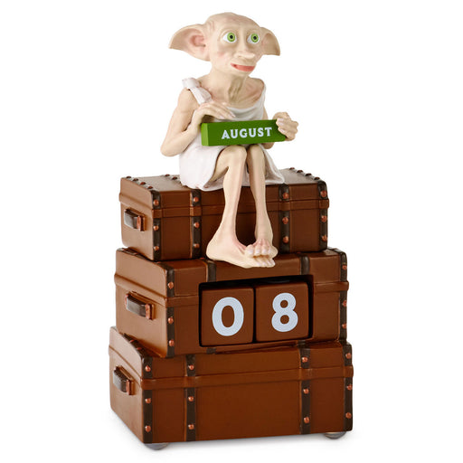 A collectible figurine of Dobby the House-Elf sitting on a stack of three brown leather-like suitcases, holding a green calendar block that reads AUGUST. The middle suitcase has flip-top blocks showing the number 08.