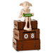 A collectible figurine of Dobby the House-Elf sitting on a stack of three brown leather-like suitcases, holding a green calendar block that reads AUGUST. The middle suitcase has flip-top blocks showing the number 08.