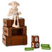 A collectible figurine of Dobby the dragon sitting on top of a stack of three brown storage trunks, with three wooden calendar blocks showing 18th February and two green blocks reading August and December, on a white background.