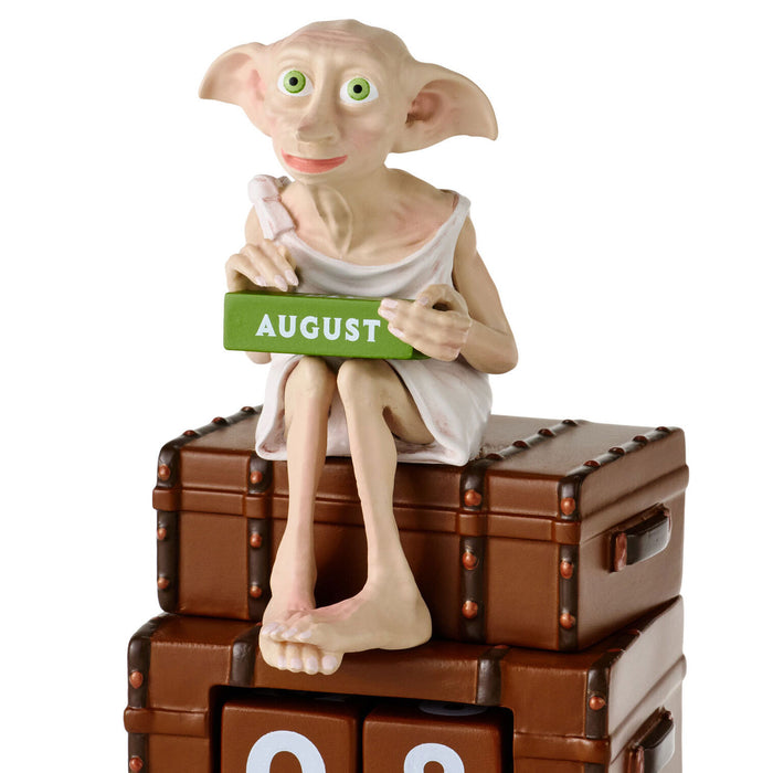 A collectible figurine of Dobby the House-elf sitting on top of two brown leather trunks, holding a green calendar block that reads 'AUGUST'.