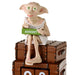 A collectible figurine of Dobby the House-elf sitting on top of two brown leather trunks, holding a green calendar block that reads 'AUGUST'.