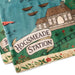A close-up of a printed textile featuring a whimsical illustration of Hogsmeade Station from Harry Potter, with a teal sky, green ground, brown house, boats, and a cream banner reading 'HOGSMEADE STATION'.