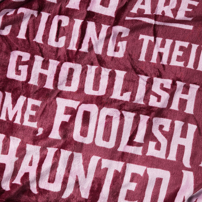 A close-up of a textured fabric, likely a throw blanket or towel, featuring a maroon background with a pale pink printed text pattern that reads 'IT'S A GHOULISH HOME, FOOLISH HAUNTED'.