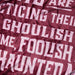 A close-up of a textured fabric, likely a throw blanket or towel, featuring a maroon background with a pale pink printed text pattern that reads 'IT'S A GHOULISH HOME, FOOLISH HAUNTED'.