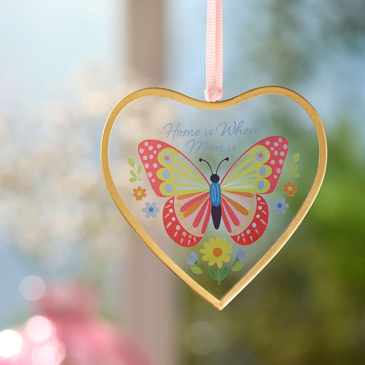 Heart-shaped decorative hanging with a butterfly design and 'Home is Where Mom is' text, against a blurred natural background.
