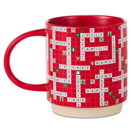 A red ceramic mug featuring a Scrabble-themed holiday design with white and green tiles spelling out Christmas words on a red grid background, set against a plain white studio background.