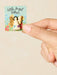 Hand holding a small angel token with a colorful card featuring flowers and text.