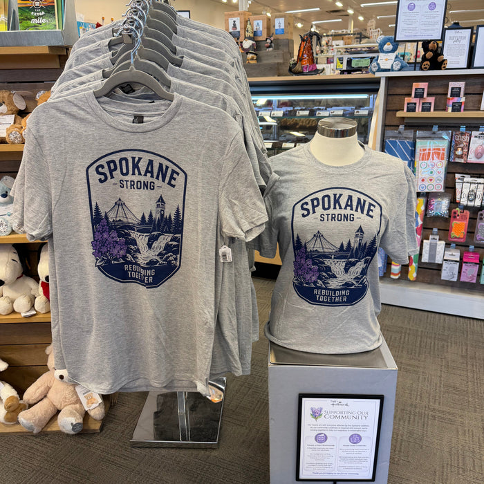 Gray graphic t-shirts displayed on hangers and a mannequin torso inside a retail store. The front shirt shows a large navy blue 'SPOKANE STRONG' shield-style graphic with a mountain and bridge illustration.