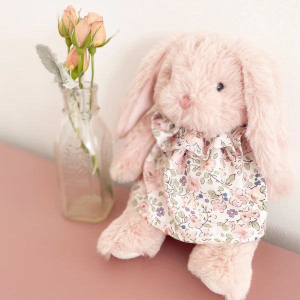 Fluffy pink bunny toy wearing a floral dress next to a small vase with flowers on a pink surface.