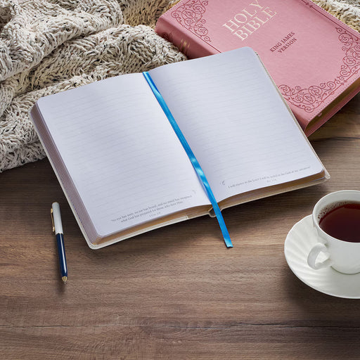 Open notebook with a pen, closed Bible, and cup of tea on a wooden surface.