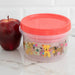 Container with a red lid and animal design next to an apple on a white surface