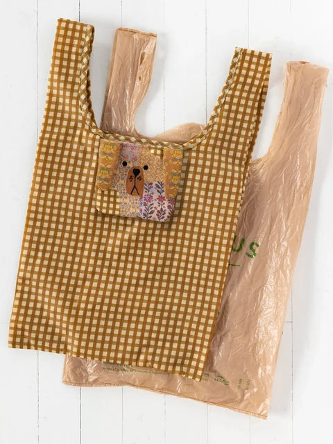 A reusable plastic shopping bag with a brown and beige gingham pattern, featuring a front pocket with a colorful dog patchwork design, shown alongside a clear plastic bag on a white wooden background.