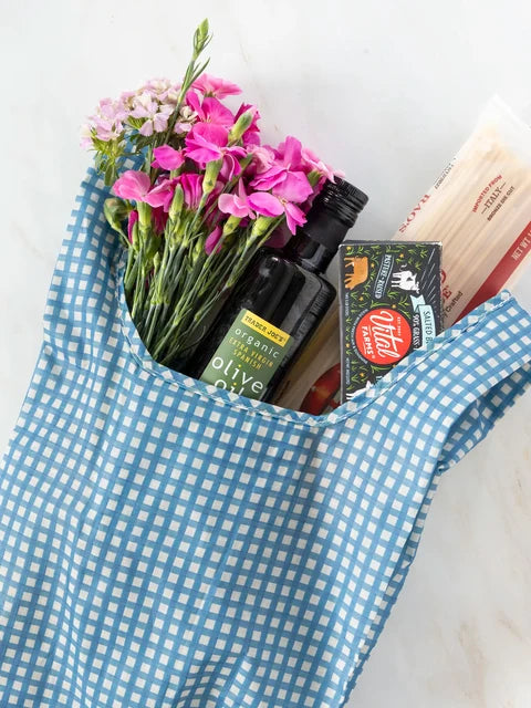 A blue and white gingham tote bag filled with pink flowers, a bottle of olive oil, a box of Oltar pasta, and a loaf of bread, set against a light marble background.