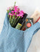 A blue and white gingham tote bag filled with pink flowers, a bottle of olive oil, a box of Oltar pasta, and a loaf of bread, set against a light marble background.