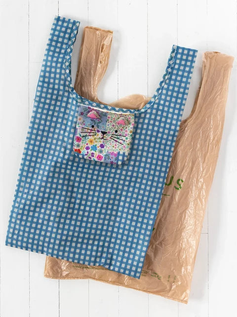 A reusable tote bag with a blue and white gingham pattern and a floral cat-face patch pocket, shown alongside a brown plastic bag on a white wood background.