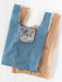 A reusable tote bag with a blue and white gingham pattern and a floral cat-face patch pocket, shown alongside a brown plastic bag on a white wood background.