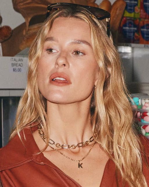 A close-up lifestyle shot of a woman with blonde hair wearing layered gold necklaces, one with a 'K' initial pendant, in what appears to be a bakery setting.