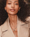 A close-up lifestyle shot of a woman with curly dark hair wearing layered silver necklaces, including a silver 'M' initial pendant, along with silver hoop earrings and a beige trench coat.