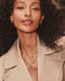 A close-up lifestyle shot of a woman with curly dark hair wearing a beige trench coat and layered silver necklaces, including a silver 'M' pendant.