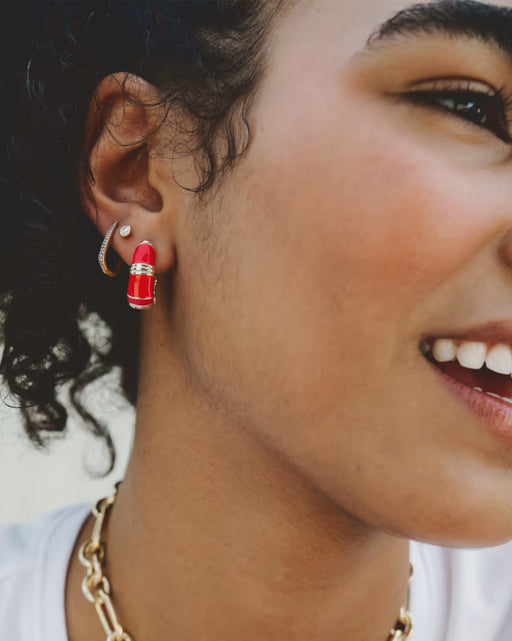 A close-up lifestyle shot of a woman wearing a pair of lipstick-shaped hoop earrings in vibrant red with gold trim, along with a gold chain necklace.