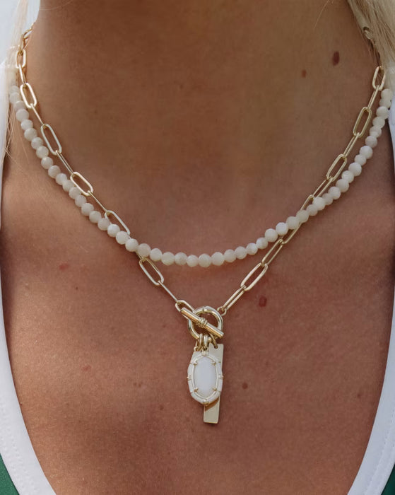 A close-up of a person wearing a layered necklace with a double strand of white seed beads and gold paperclip links, featuring a gold toggle clasp and an oval white pendant.