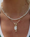 A close-up of a person wearing a layered necklace with a double strand of white seed beads and gold paperclip links, featuring a gold toggle clasp and an oval white pendant.