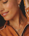 Close-up of a woman wearing layered necklaces, including a silver-toned longhorn pendant necklace and delicate star chain necklaces, paired with an orange-brown collared shirt.