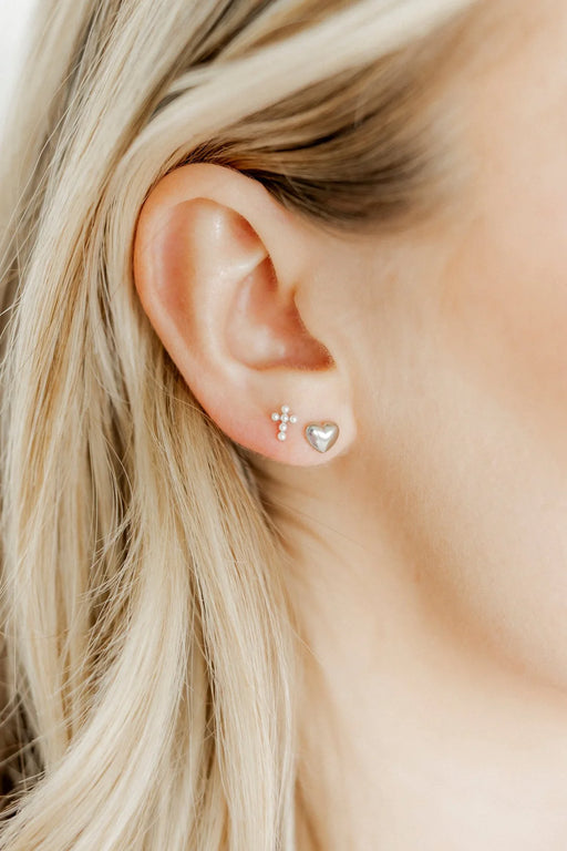 Close-up of an ear wearing gold stud earrings with a cross and heart design.
