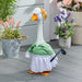A decorative goose figurine dressed as a golf player, wearing a green polo shirt and white skirt, holding a golf club and standing on a gray wood floor with potted plants in the background.