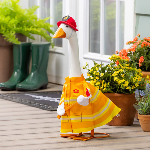 Decorative goose in a yellow raincoat and red hat on a wooden deck with plants and boots in the background.