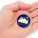 A circular lapel pin featuring a white paddle steamer ship on a dark blue background with a gold border, held in the palm of a hand.