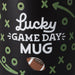 Black mug with 'Lucky Game Day Mug' text and football design on a black background with green symbols.