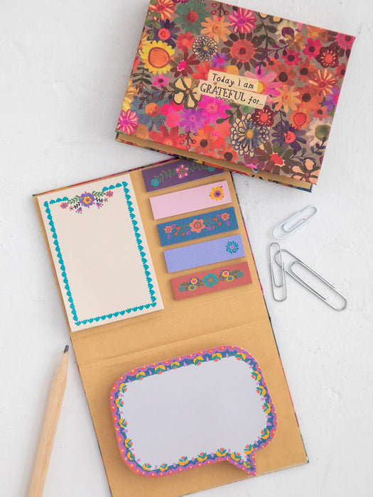 A floral stationery set displayed on a light surface, including a patterned box lid with a handwritten note, an open kraft paper folder with matching floral notepad pages, decorative sticky note tabs, and metal paperclips.