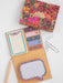 A floral stationery set displayed on a light surface, including a patterned box lid with a handwritten note, an open kraft paper folder with matching floral notepad pages, decorative sticky note tabs, and metal paperclips.