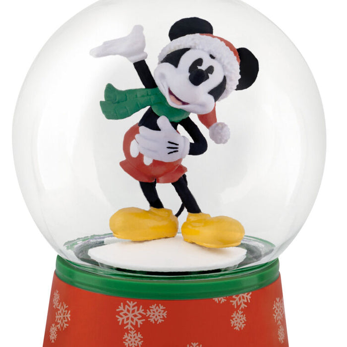 A Disney Mickey Mouse snow globe figurine featuring Mickey dressed in a Santa hat, green scarf, red shorts, and yellow shoes, set inside a clear glass globe on a red and green patterned base.