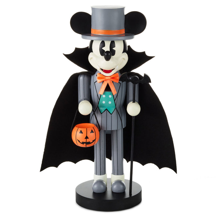 A Mickey Mouse Halloween nutcracker figurine dressed as a bat, wearing a gray pinstripe suit, black cape, gray top hat with an orange band, orange bow tie, green polka-dot tie, and holding an orange jack-o'-lantern bucket, standing on a black base against a white background.
