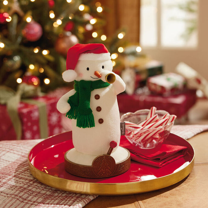 Decorative snowman figurine with a red hat and green scarf on a table with a Christmas tree in the background.