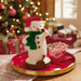 Decorative snowman figurine with a red hat and green scarf on a table with a Christmas tree in the background.