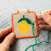 Hand holding a cross-stitched pineapple patch with green thread being sewn on a gray fabric background.