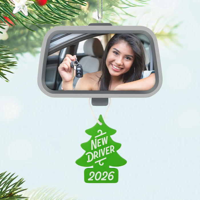 Car mirror with a reflection of a woman holding keys, Christmas tree ornament labeled 'New Driver 2026' on a light blue background.
