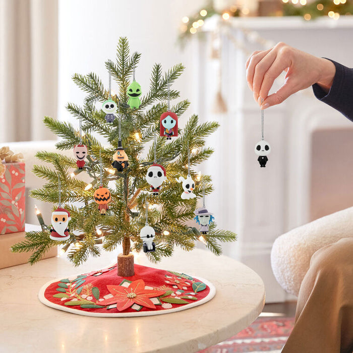 A small decorated Christmas tree on a round table in a cozy living room, with a hand hanging a small character ornament from the tree. The tree has green needles, warm white string lights, and several colorful character ornaments, and the base is covered by a red holiday tree skirt.