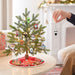 A small decorated Christmas tree on a round table in a cozy living room, with a hand hanging a small character ornament from the tree. The tree has green needles, warm white string lights, and several colorful character ornaments, and the base is covered by a red holiday tree skirt.