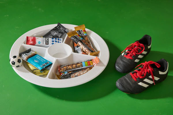 Snack tray with various bars and a soccer ball next to soccer cleats on a green background