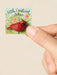 A hand holds a small square card featuring a red cardinal figurine and the text 'Little Cardinal token' against a plain beige background.