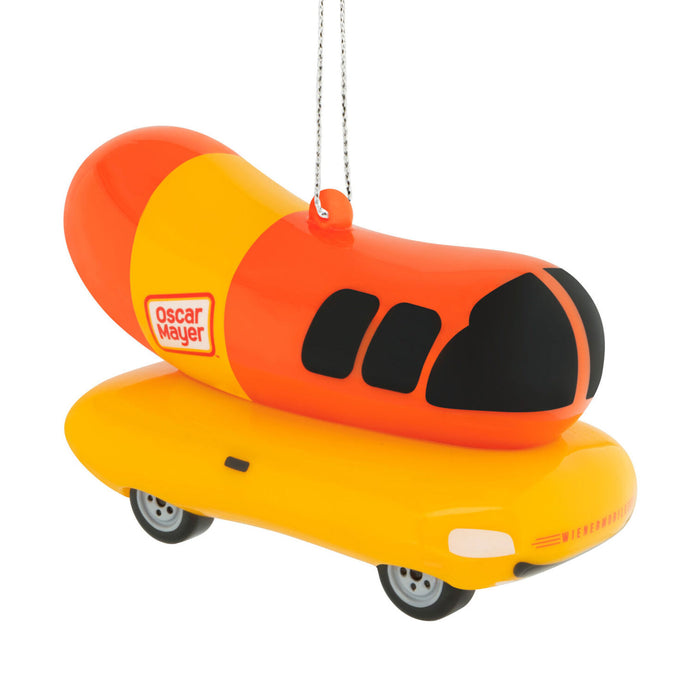 An Oscar Mayer hot dog ornament featuring a bright orange and yellow hot dog body with black eyes, mounted on a yellow car base with small wheels, hanging by a silver string against a white background.