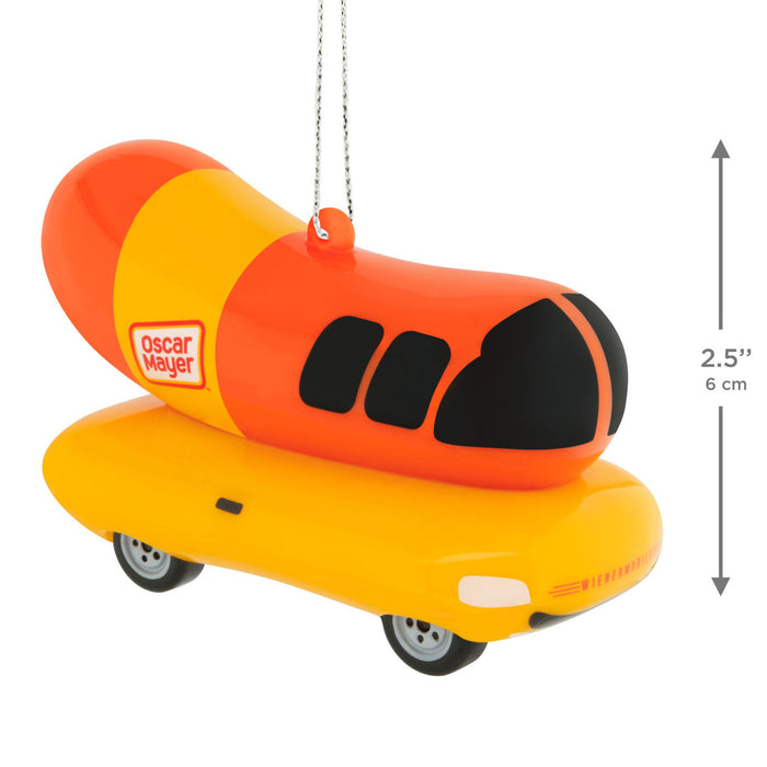 An Oscar Mayer hot dog ornament featuring a bright orange and yellow hot dog body with black eyes, mounted on a yellow car base with hanging wires, shown on a white background with a height measurement annotation.