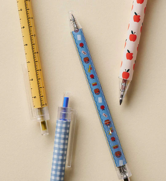 Three novelty school supply pens arranged on a light beige background: a yellow ruler pen, a blue gingham pencil sharpener pen, and a white pen with red apple illustrations.