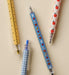 Three novelty school supply pens arranged on a light beige background: a yellow ruler pen, a blue gingham pencil sharpener pen, and a white pen with red apple illustrations.