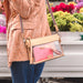 A person wearing a textured peach jacket and blue jeans carries a clear plastic crossbody bag with a beige frame and a pink glittery wallet inside. The bag has a rainbow-striped shoulder strap.